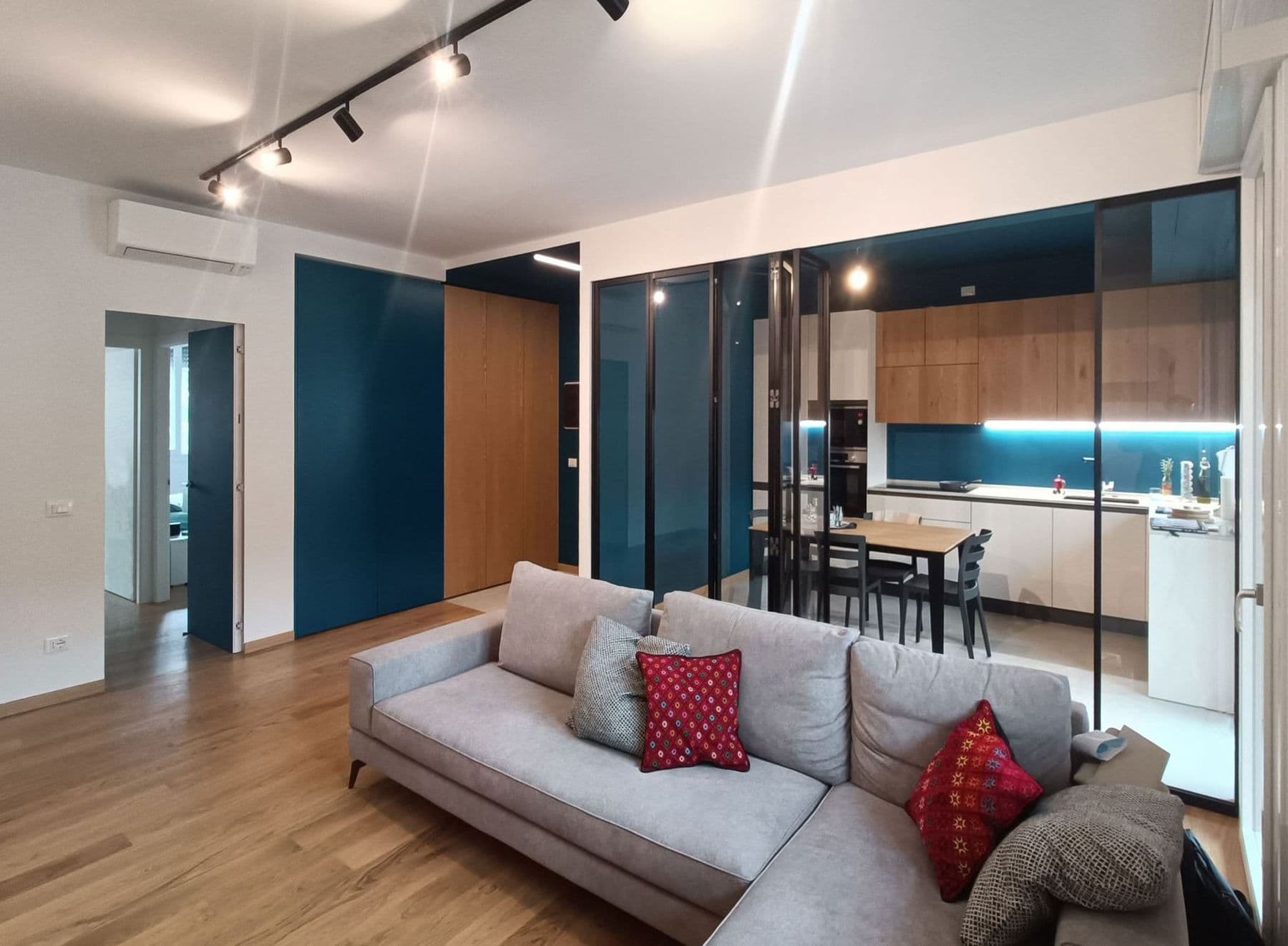 Casa Vito, Ferrara: soggiorno open space con divano grigio, parquet in rovere e cucina blu petrolio dietro una vetrata