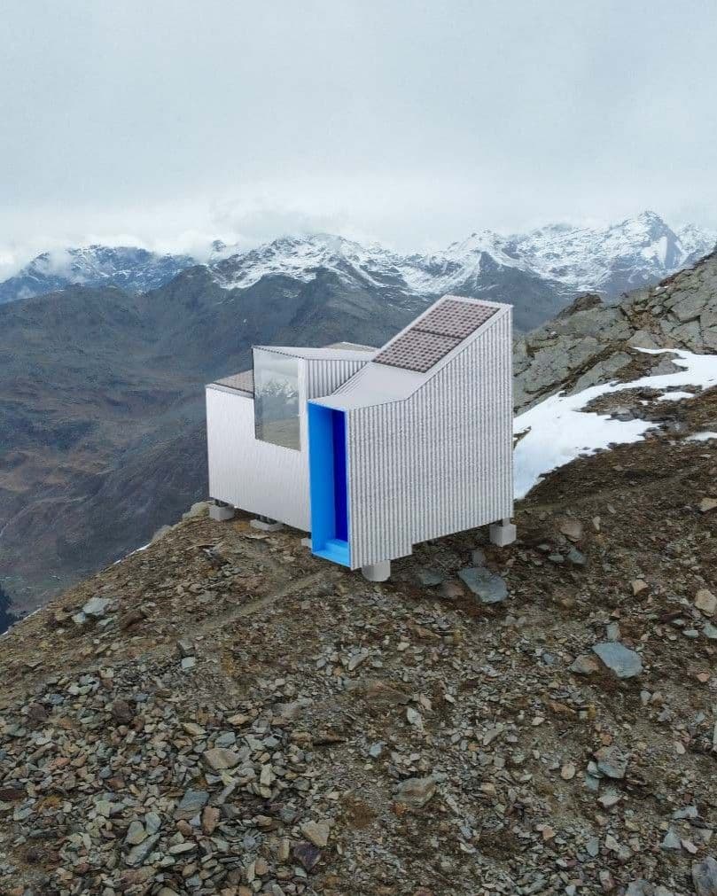 Mountain Guardian: vista ravvicinata del bivacco con l’ingresso rivestito di blu e le cime innevate sullo sfondo